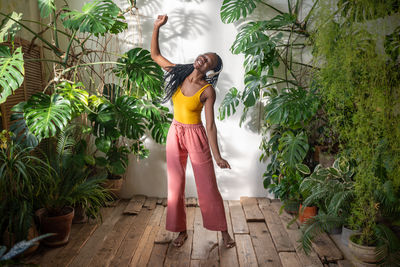 Happy carefree black woman dancing with closed eyes listening music in headphones among houseplants