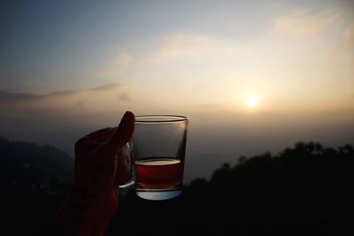 Close-up of hand holding beer glass against sky during sunset