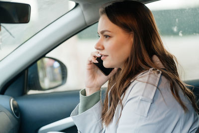 Beautiful girl with long hair in a grey trench coat using smartphone call gets into the car