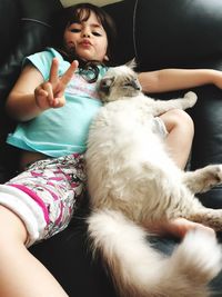 Portrait of girl gesturing peace sign while lying with cat on sofa