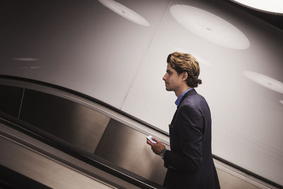 Side view of businessman holding mobile phone on escalator in airport