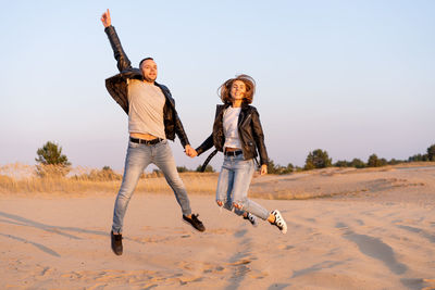 Couple holding hands and jumping at beach against clear sky