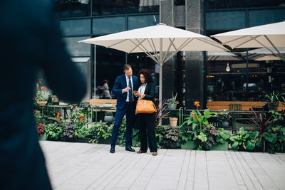 Full length of businessman and businesswoman sharing mobile phone against parasol in city
