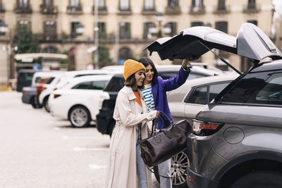 Positive female friends in stylish wear putting bag into truck of modern automobile parked on street of city with building