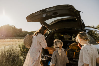 Mother unloading basket from car trunk with boys by meadow at sunny day