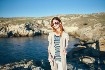 Young woman wearing sunglasses standing on rock against sky