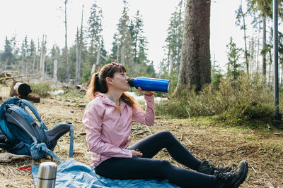 Young beautiful woman drinking hot tea or coffee from a thermos in the forest on a walk. hiking