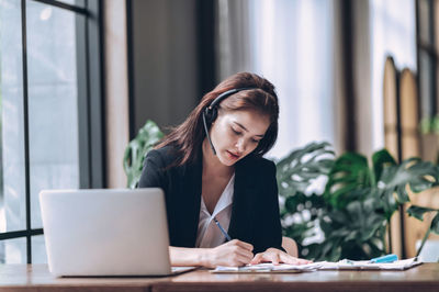 Businesswomen in headphones writing notes in notebook and watching webinar online