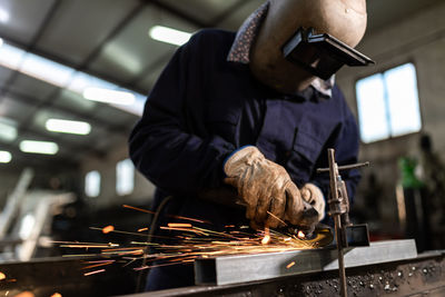 Unrecognizable male employee in protective gloves and helmet using welding machine while working in dark workshop