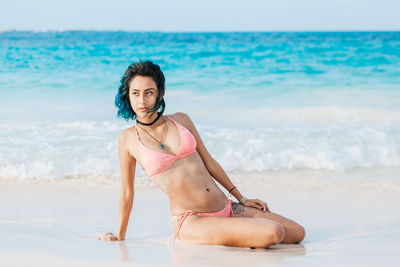 Woman in bikini kneeling on shore at beach against sky