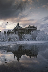 Fountain in lake against buildings during sunset