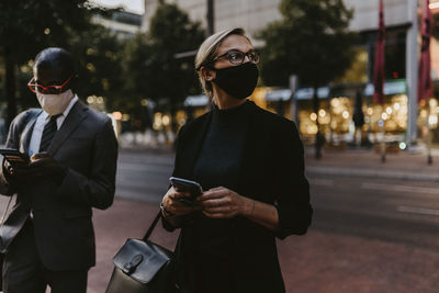 Female entrepreneur with smart phone looking away while standing by male colleague during covid-19