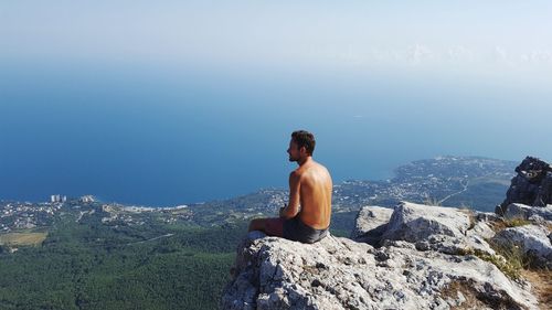 Shirtless man looking at sea while sitting on cliff during sunny day