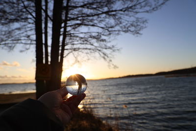 Cropped hand of person holding crystal ball at beach against sky during sunset