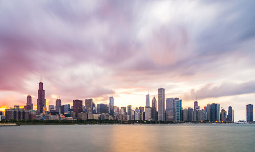 Sea by modern buildings against sky during sunset