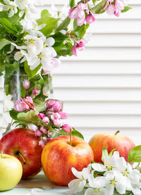 Apples close up and apple tree blossom branches in glass vase on white window blind backdrop.