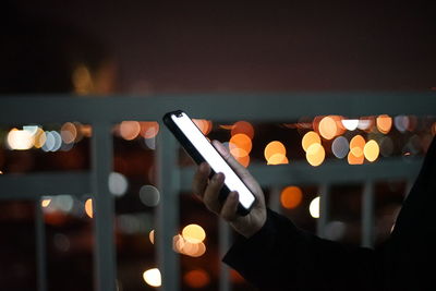 Cropped hand holding mobile phone against defocused lights at night