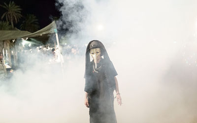 Man wearing mask while standing amidst smoke at night