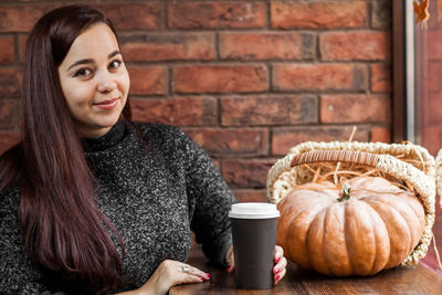 Portrait of woman sitting with disposable cup and pumpkin against brick wall