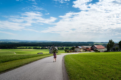 Rear view of woman walking on road amidst grassy field against cloudy sky