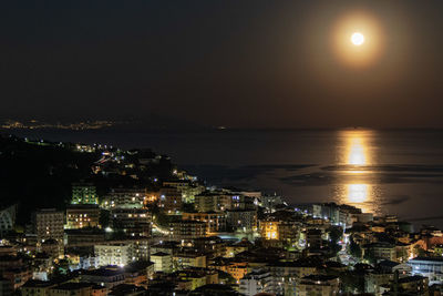 High angle view of illuminated city by sea against sky at night