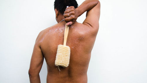Rear view of shirtless man rubbing back with brush while standing against wall