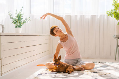 Beautiful adult woman practicing yoga pose with pygmy pinscher dog enjoying