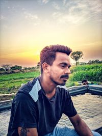 Portrait of young man looking away while sitting against sky during sunset