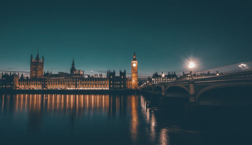 Illuminated bridge over river by buildings against sky at night