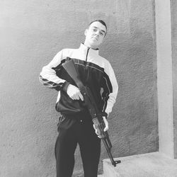 Portrait of young man holding rifle while standing against wall