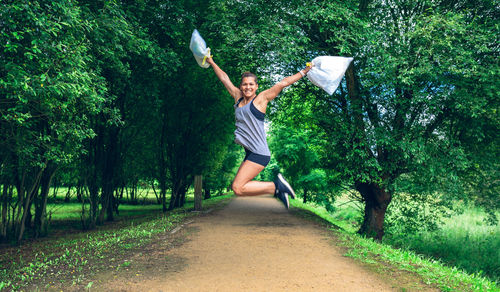 Portrait of cheerful woman holding garbage in plastic bags while jumping at park