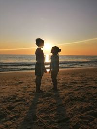 Siblings standing on beach against sky during sunset