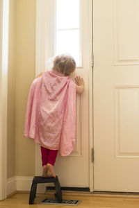 Sweet blonde girl wearing pink cape stands on stool, looks out window