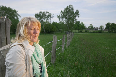 Lateral torso view of a mature woman standing by a fence and gazing to the right into the light.