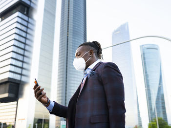 Side view of determined african american male entrepreneur in protective mask in downtown during coronavirus browsing smartphone on street