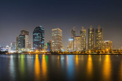 Illuminated modern buildings by bay against sky at night