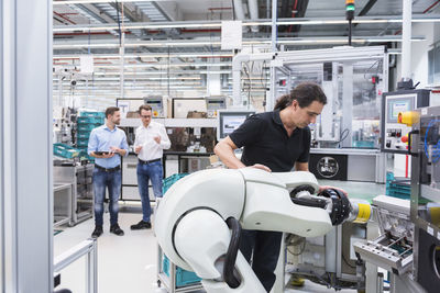 Man operating assembly robot in factory with two men in background supervising