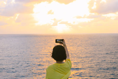 Rear view of man photographing sea against sky during sunset