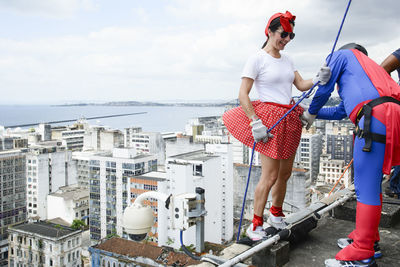 Woman wearing hero costume being prepared by a man to descend a tall rappel building. 