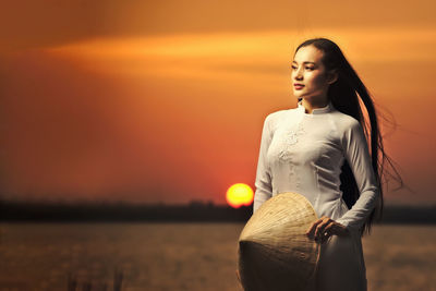 Young woman looking away while standing against orange sunset sky