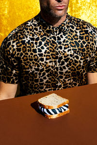 Crop anonymous male in shirt with leopard print sitting at brown table with sandwich with zebra filling in light room