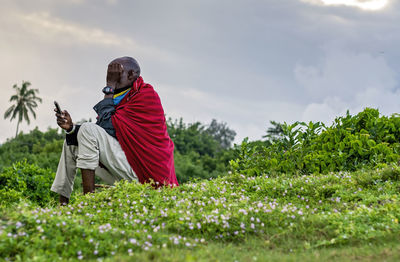 Maasai man sitting on a hill using his mobile phone against sky