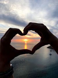 Cropped hands making heart shape at beach against sky during sunset
