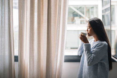 Side view of young woman drinking coffee at window