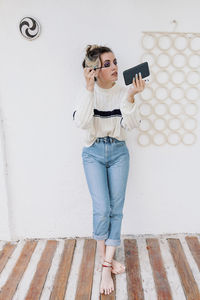 Beautiful woman applying mascara looking in smart phone while standing against white wall