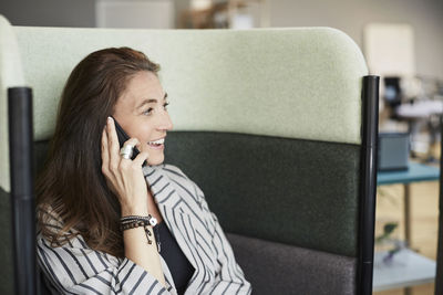 Smiling mature businesswoman talking through mobile phone while sitting on chair at creative office