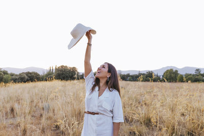 Smiling woman holding hat while standing by plants on land against clear sky