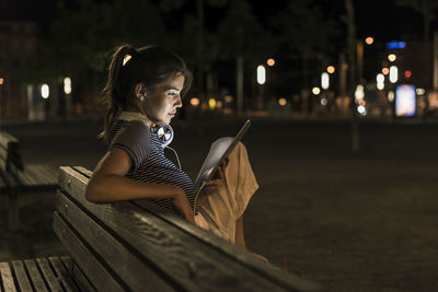 Young woman sitting on bench at night using tablet