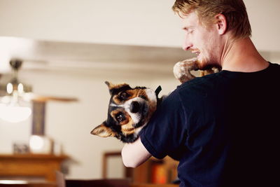 Rear view of young man carrying dog while standing at home