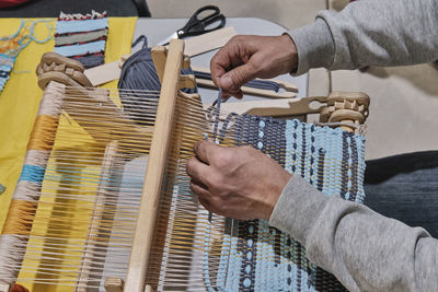 Hands of senior man weaving small rug with pattern on manual table loom.